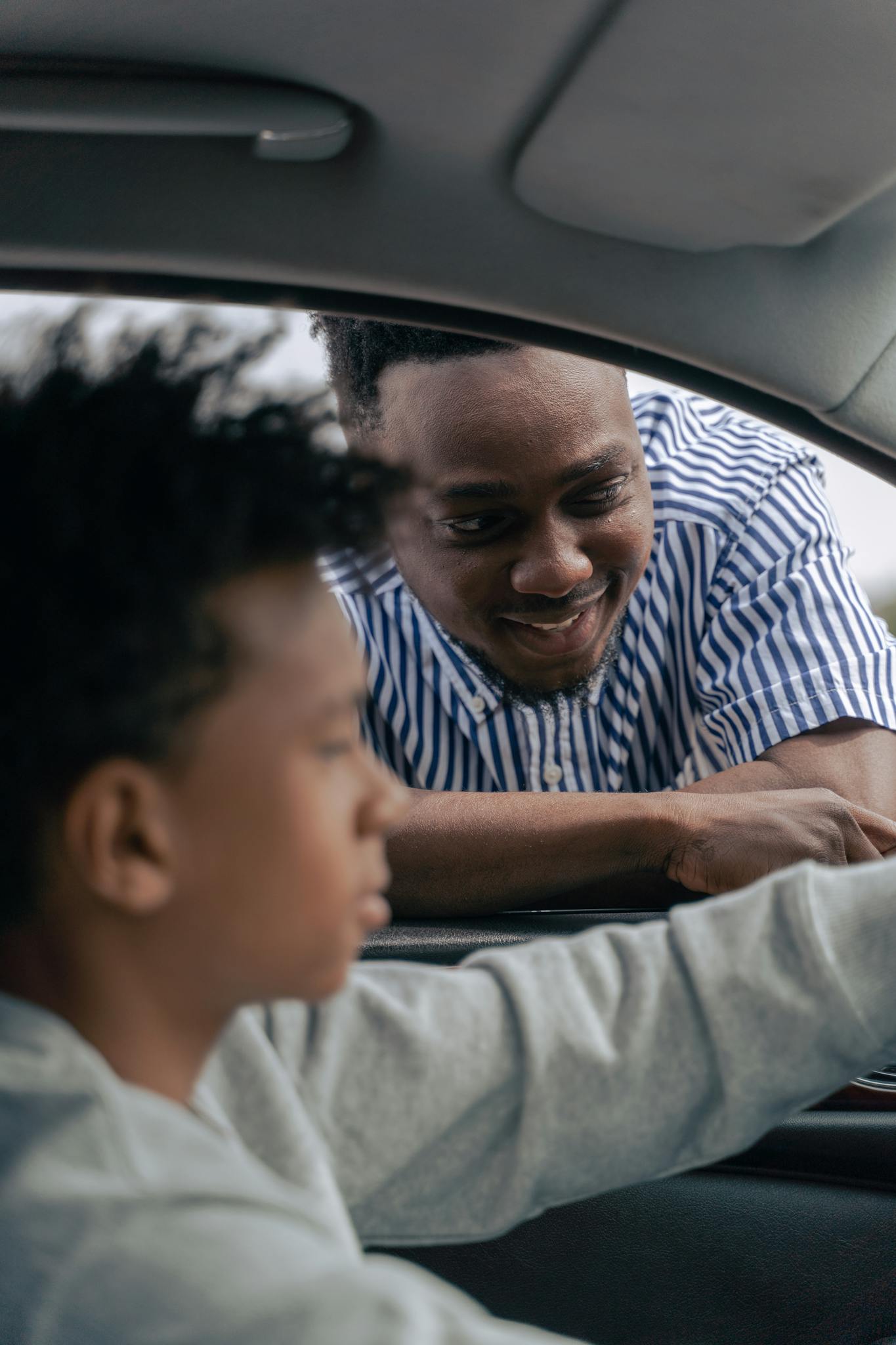 A man smiles as his son pretends to drive a car indoors, showcasing a special family moment.