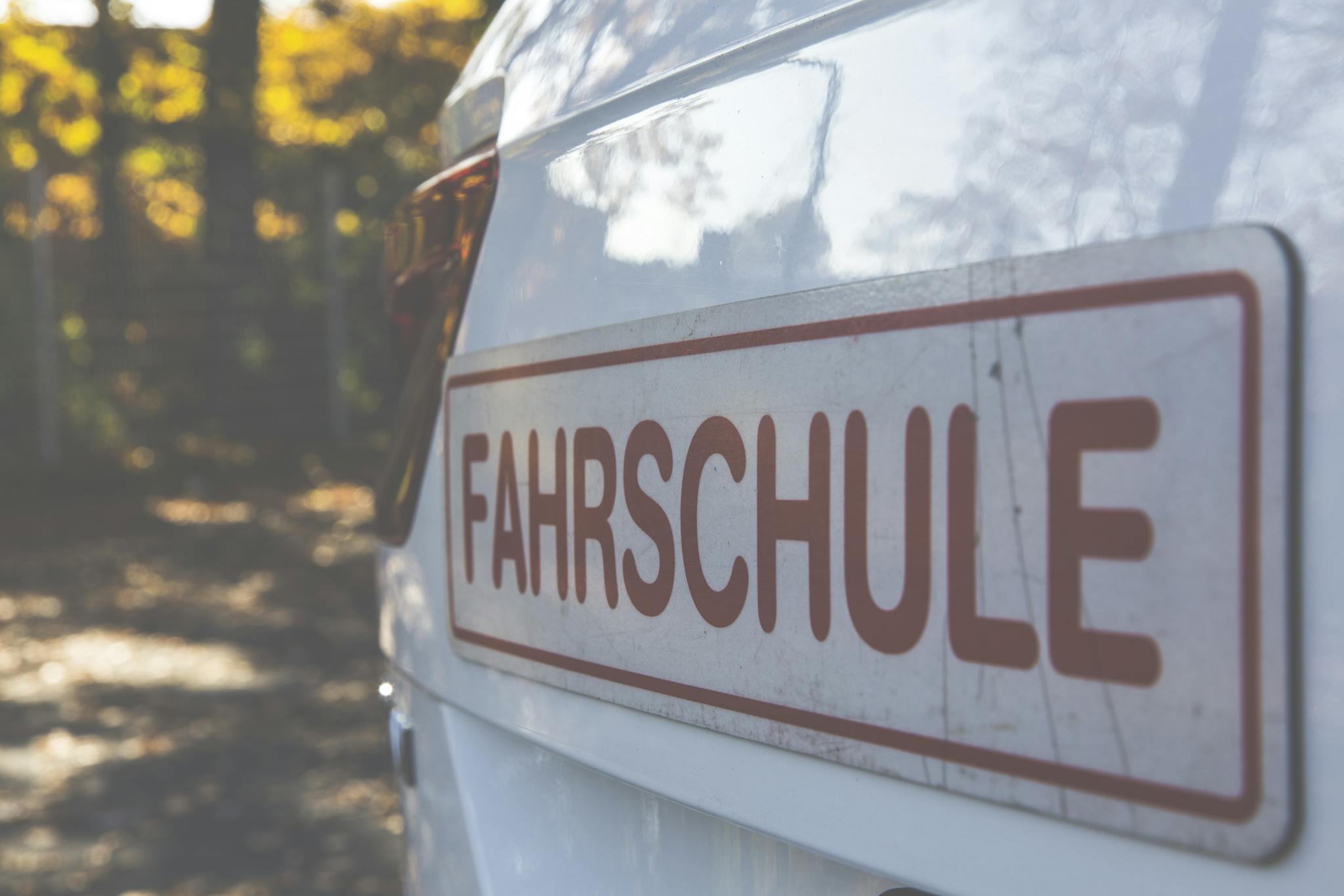 A side view of a white car with a Fahrschule sign, outdoors with autumn foliage.