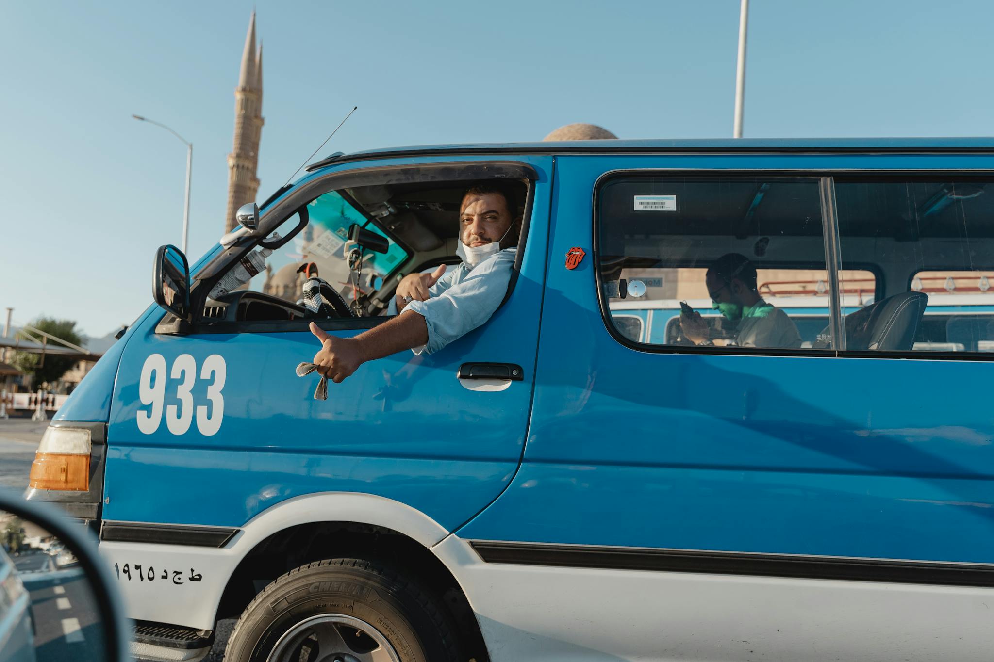 Driver with facemask in blue van on busy urban road during daylight.